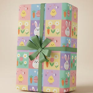 Peut inclure: Un coffret cadeau emballé dans du papier à motifs de Pâques. Le motif répété présente des poussins, des lapins, des carottes et des fleurs. Le coffret est noué avec un ruban et un nœud vert.