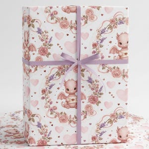 May include: A gift box wrapped in paper with a repeating pattern of pink dragons, roses, hearts, and lavender sprigs on a white background. The box is tied with a lavender ribbon. The wrapping paper is also visible.