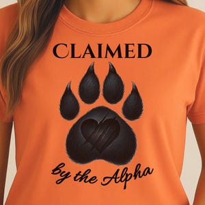 May include: Orange t-shirt featuring a black paw print design with a heart and claw marks. The text "CLAIMED" is above the paw print, and "by the Alpha" is below it, all in black script.