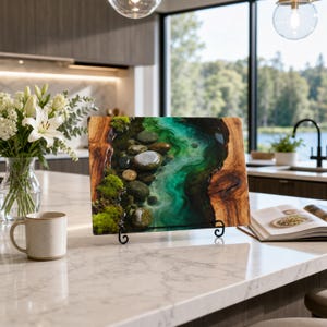 May include: A rectangular wooden serving board featuring a central resin inlay that mimics a flowing river with vibrant green and turquoise hues, embedded with smooth river stones and patches of green moss, displayed on a black metal stand.