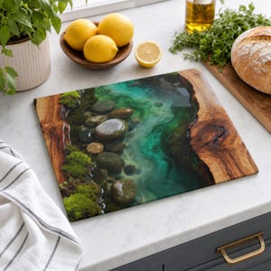 May include: A rectangular chopping board featuring a natural wood edge and a central resin inlay depicting a clear turquoise river stream with smooth grey river stones and patches of vibrant green moss along the bank.