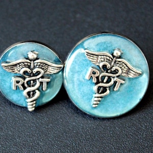 May include: Two round, blue and silver medical symbol earrings. Each earring features a silver caduceus symbol with the letters "ROT" in silver. The background is a light blue color. The earrings are displayed on a dark surface.