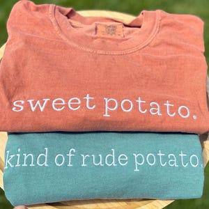 May include: Two t-shirts, one coral and one teal, are stacked on top of each other. The coral shirt has the embroidered text "sweet potato." The teal shirt has the embroidered text "kind of rude potato."