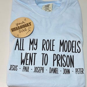 May include: Light blue Comfort Colors t-shirt with the text "ALL MY ROLE MODELS WENT TO PRISON" in black. Below the text are the names Jesus, Paul, Joseph, Daniel, John, and Peter. A wooden tag with the text "your EMBROIDERY Girl" is on the shirt.
