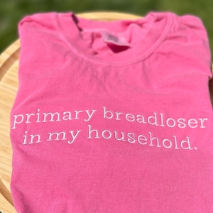 May include: A folded, bright pink t-shirt with the embroidered text "primary breadloser in my household." The shirt is made of a soft material and is displayed on a wooden surface.