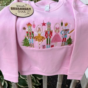 May include: Pink sweatshirt with an embroidered Nutcracker design. The design features several characters, including a nutcracker soldier, a ballerina, and a mouse king, along with Christmas trees and stars. A wooden tag with the text "Your Embroidery Girl" is attached.