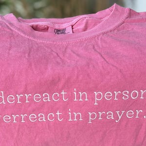 May include: A pink short-sleeved t-shirt with a crew neckline. The shirt features the white text "underreact in person, overreact in prayer." printed on the front. The interior neck label displays the brand name Comfort Colors in black text.