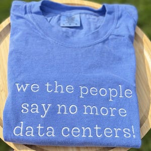 May include: A light blue t-shirt with the embroidered text "we the people say no more data centers!". The shirt is folded and appears to be made of a soft, comfortable material. The shirt is on a wooden surface.