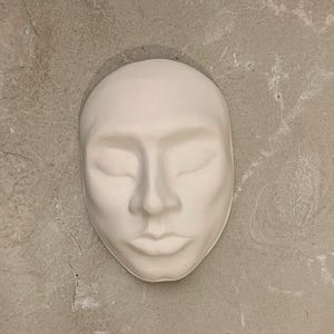 May include: A white plaster face mask with closed eyes, a defined nose, and full lips. The mask is set against a textured, light gray background. The mask is a decorative item.