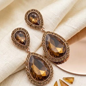 May include: A pair of elegant drop earrings with large, teardrop-shaped amber-colored stones. The stones are surrounded by a halo of smaller, matching crystals, set in a rose-gold tone metal. Additional loose crystals are scattered nearby.