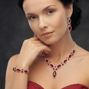 May include: A jewelry set consisting of a necklace, a bracelet, and drop earrings. Each piece features marquise-cut red stones surrounded by small clear crystals set in a silver-toned metal frame. The necklace has a central marquise-cut pendant.
