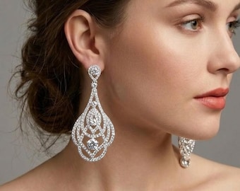 May include: A pair of silver-toned chandelier earrings featuring a teardrop-shaped filigree design encrusted with clear, sparkling crystals. Each earring is anchored by a single round crystal stud at the top, leading to an intricate lattice pattern.