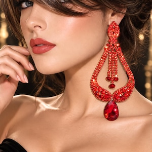 May include: A large, chandelier-style earring featuring a teardrop-shaped frame encrusted with numerous small, vibrant red crystals. The design includes three dangling crystal strands in the center and a single, prominent red teardrop gem at the base.