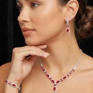 May include: A jewelry set featuring a necklace, bracelet, and earrings. The set includes red marquise-cut stones and clear round stones set in a silver-toned metal. The necklace and bracelet are connected by a chain.
