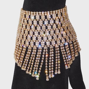May include: A gold-toned rhinestone cuff bracelet. The bracelet features a grid-like design with dangling rhinestone fringe. The jewelry is displayed on a black surface, creating a contrast that highlights the sparkle of the rhinestones.