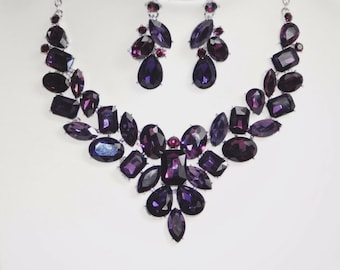 May include: A matching necklace and earring set featuring large, faceted, dark purple gemstones. The necklace has a V-shaped design with various shapes of stones. The earrings are teardrop-shaped with smaller stones.