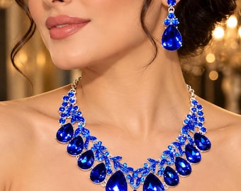 May include: A silver-toned necklace and matching drop earrings set featuring large, faceted, pear-shaped royal blue crystals. The necklace is composed of multiple teardrop-shaped blue stones arranged in a cluster, accented by smaller round blue crystals.