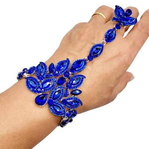 May include: A blue hand chain bracelet with a floral design. The bracelet features multiple blue gemstones in a leaf-like shape, connected by a silver chain. A ring with a blue flower is attached to the chain.