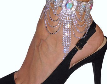 May include: A silver anklet with cascading crystal chains and large, iridescent, oval-shaped gems. The anklet is worn with a black high-heeled shoe, creating a glamorous look. The crystals sparkle, reflecting light.
