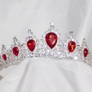 May include: A silver-toned tiara with sparkling clear crystals and vibrant red teardrop-shaped gemstones. The crown is set against a white background, showcasing its intricate design and elegant details. A regal accessory.