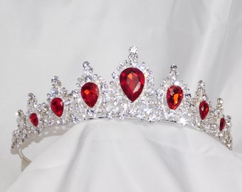 May include: A silver-toned tiara with sparkling clear crystals and vibrant red teardrop-shaped gemstones. The crown is set against a white background, showcasing its intricate design and elegant details. A regal accessory.