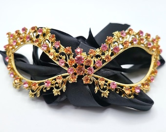May include: A gold-coloured masquerade mask with pink and orange gemstones. The mask features a floral design and is attached to a black satin ribbon. Ideal for a formal event or costume party.
