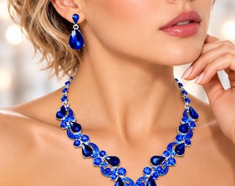 May include: A silver-toned necklace and matching drop earring set featuring deep blue teardrop-shaped stones and smaller round blue crystals arranged in a V-shaped pattern. The jewelry set is crafted with a metallic silver finish.