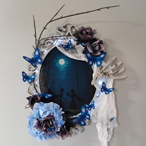 May include: Oval wall art featuring a dark forest scene with a full moon. The frame is decorated with blue butterflies, artificial flowers in shades of blue and purple, and a skeletal hand. Twigs and fairy lights add to the whimsical design.