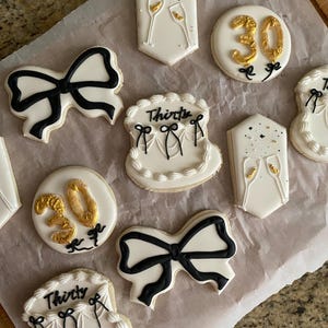 May include: Assortment of decorated sugar cookies with a black and white theme. Cookies include bow shapes, champagne flutes, and the number 30 in gold. Some cookies are shaped like cakes and have the word "Thirty" on them.