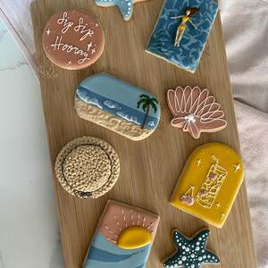 May include: A collection of eight decorated sugar cookies on a wooden board. Designs include a light blue starfish, a round cookie with the text "Sip Sip Hooray", a blue pool scene with a figure, a beach scene, a seashell, a straw hat, a drink, and a sunset.