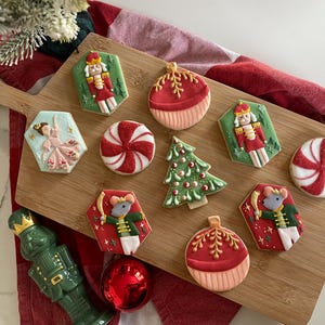 May include: A wooden cutting board displays an assortment of Christmas cookies. The cookies include nutcrackers, ornaments, a Christmas tree, a ballerina, and peppermint candies. The cookies are decorated with red, green, white, and gold icing.