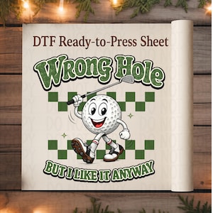 May include: A DTF ready-to-press sheet featuring a cartoon golf ball character with arms and legs, holding a golf club. The text reads "Wrong Hole" and "But I Like It Anyway" in green, with a green checkered pattern background behind the character.