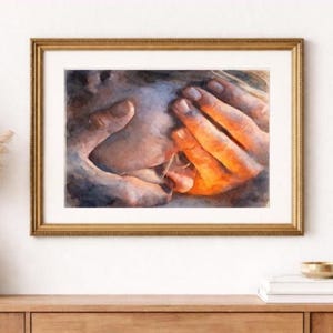 May include: Watercolor print of hands in warm tones, framed in a gold-colored frame. The artwork depicts hands gently touching, with a focus on the interplay of light and shadow. The art piece is displayed on a wooden surface.