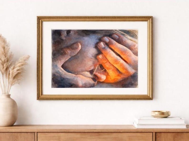 May include: Watercolor print of hands in warm tones, framed in a gold-colored frame. The artwork depicts hands gently touching, with a focus on the interplay of light and shadow. The art piece is displayed on a wooden surface.