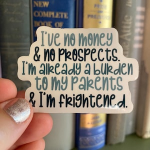 May include: A white sticker with the text "I've no money & no prospects. I'm already a burden to my parents & I'm frightened." The text is in blue and black. The sticker is held in front of a bookshelf with hardback books.