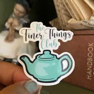 Pode incluir: Um autocolante com uma chaleira turquesa com contorno preto e o texto "The Finer Things Club". O autocolante é segurado em frente a um fundo desfocado com uma figura e um livro.