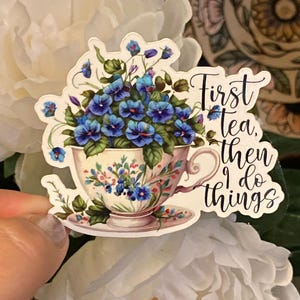 Puede incluir: Una pegatina con una taza de té rebosante de flores azules. La taza de té tiene un diseño floral y se apoya en un platillo. El texto "First tea, then I do things" está escrito en letra negra.