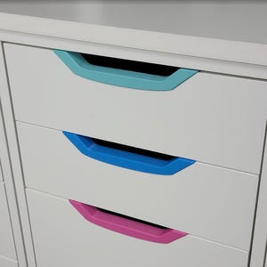 May include: A white cabinet with three drawers, each featuring a different colored handle: teal, blue, and pink. The handles are recessed and provide a pop of color against the white surface.
