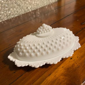 May include: White hobnail-patterned butter dish with a scalloped edge. The lid features a cluster of hobnails at the top. The dish is oval-shaped and sits on a wooden surface.