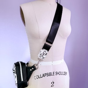 Peut inclure: Sac banane noir et blanc avec une bandouli&egrave;re assortie. La bandouli&egrave;re a une boucle d&eacute;corative avec un motif noir et blanc. Le sac banane est attach&eacute; &agrave; la bandouli&egrave;re avec un anneau en m&eacute;tal argent&eacute;.