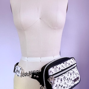 Peut inclure: Sac banane noir et blanc avec un motif Mickey Mouse, pr&eacute;sent&eacute; sur un mannequin. Le sac a une fermeture &eacute;clair noire et une cha&icirc;ne argent&eacute;e. La ceinture est blanche et le sac est attach&eacute; avec un fermoir argent&eacute;.