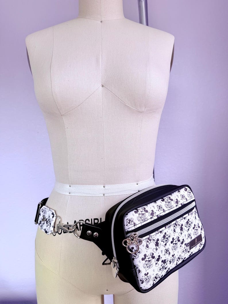 Peut inclure: Sac banane noir et blanc avec un motif Mickey Mouse, pr&eacute;sent&eacute; sur un mannequin. Le sac a une fermeture &eacute;clair noire et une cha&icirc;ne argent&eacute;e. La ceinture est blanche et le sac est attach&eacute; avec un fermoir argent&eacute;.