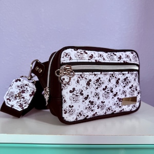 Peut inclure: Sac banane noir et blanc avec un motif Mickey Mouse. Il a une bordure marron, des fermetures &eacute;clair argent&eacute;es et une &eacute;tiquette assortie. Le sac est pos&eacute; sur une surface blanche.