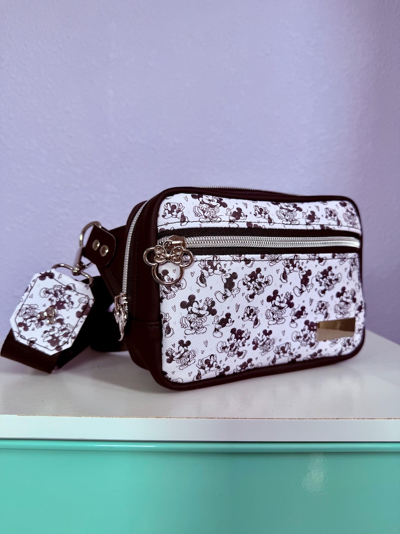 Peut inclure: Sac banane noir et blanc avec un motif Mickey Mouse. Il a une bordure marron, des fermetures &eacute;clair argent&eacute;es et une &eacute;tiquette assortie. Le sac est pos&eacute; sur une surface blanche.