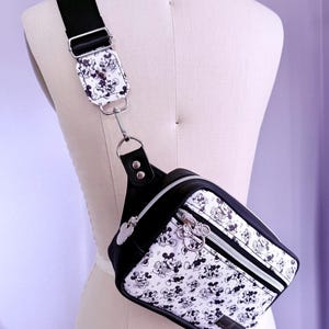 Peut inclure: Sac bandouli&egrave;re noir et blanc avec un motif Mickey Mouse r&eacute;p&eacute;t&eacute;. Le sac est dot&eacute; d'une bandouli&egrave;re et d'une garniture noires, d'une fermeture &eacute;clair argent&eacute;e et d'une tirette Mickey Mouse argent&eacute;e. Le sac est pr&eacute;sent&eacute; sur un mannequin.
