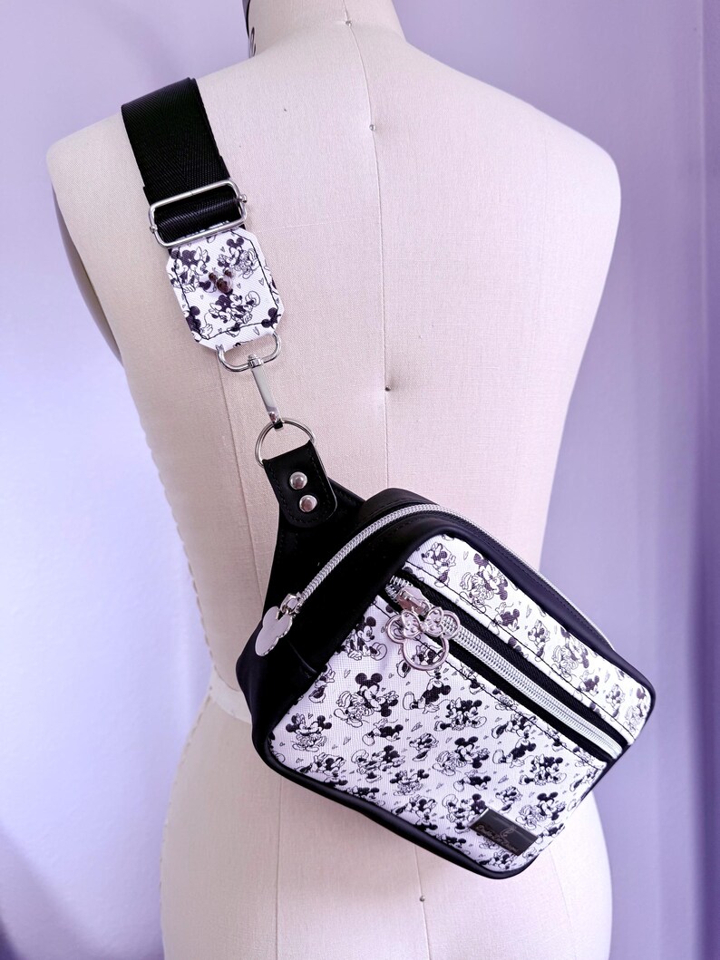 Peut inclure: Sac bandouli&egrave;re noir et blanc avec un motif Mickey Mouse r&eacute;p&eacute;t&eacute;. Le sac est dot&eacute; d'une bandouli&egrave;re et d'une garniture noires, d'une fermeture &eacute;clair argent&eacute;e et d'une tirette Mickey Mouse argent&eacute;e. Le sac est pr&eacute;sent&eacute; sur un mannequin.