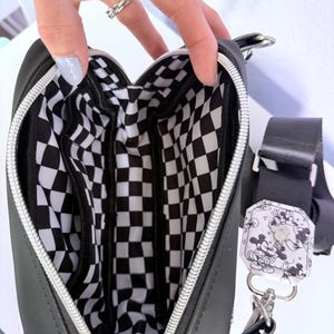 Peut inclure: Sac bandouli&egrave;re noir avec une fermeture &eacute;clair argent&eacute;e et un int&eacute;rieur &agrave; carreaux noir et blanc. Le sac a une sangle noire et une petite &eacute;tiquette d&eacute;corative avec un motif Mickey Mouse. Le sac est ouvert, r&eacute;v&eacute;lant plusieurs compartiments.
