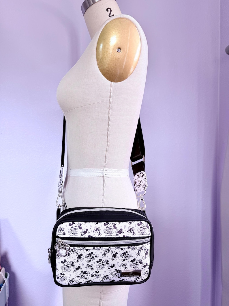 Peut inclure: Sac bandouli&egrave;re noir et blanc avec un motif Mickey Mouse r&eacute;p&eacute;t&eacute;. Le sac a un dessus et un dessous noirs, des accessoires argent&eacute;s et une bandouli&egrave;re noire. Le sac est pr&eacute;sent&eacute; sur un mannequin.
