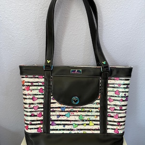 May include: A black tote bag with a white and black striped pattern, decorated with colorful heart shapes and paint splatters. The bag features black handles, a front pocket, and a decorative clasp with a character icon.