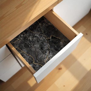 May include: A drawer with a black marble-patterned liner featuring gold veining. The drawer is open, revealing the decorative liner inside a wooden frame with a white front. The surrounding furniture is light-colored wood.